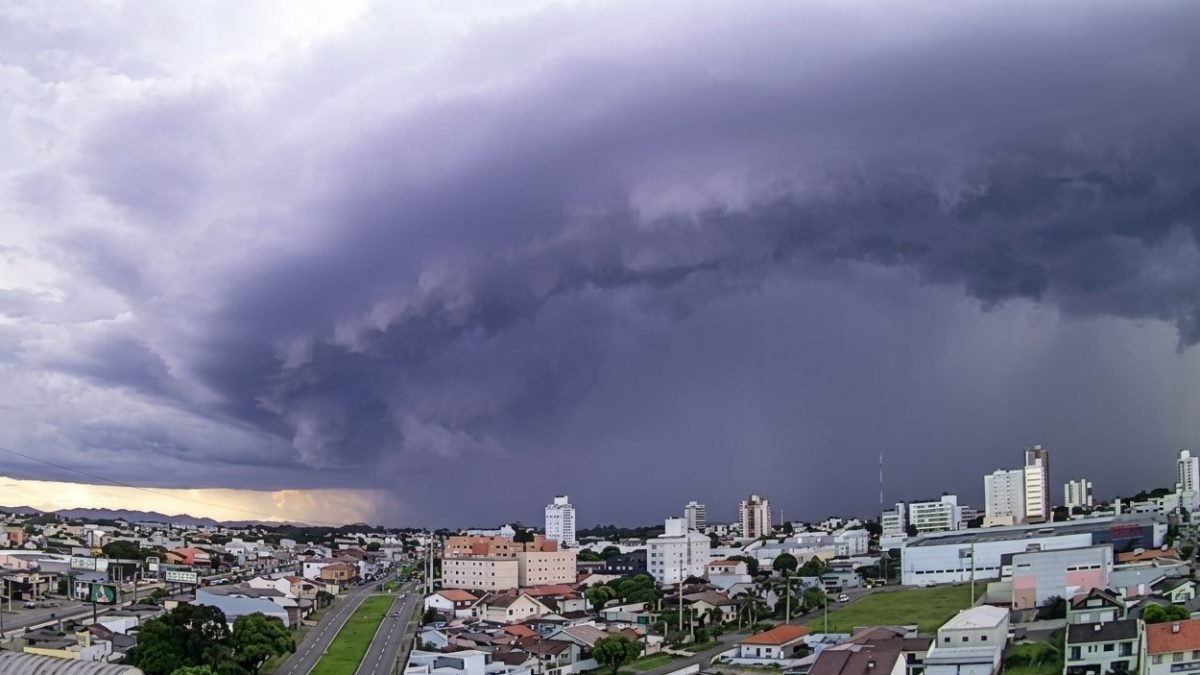 Dark shelf cloud over a city signals an approaching storm, with rain visible in the distance and low sunlight on the horizon.