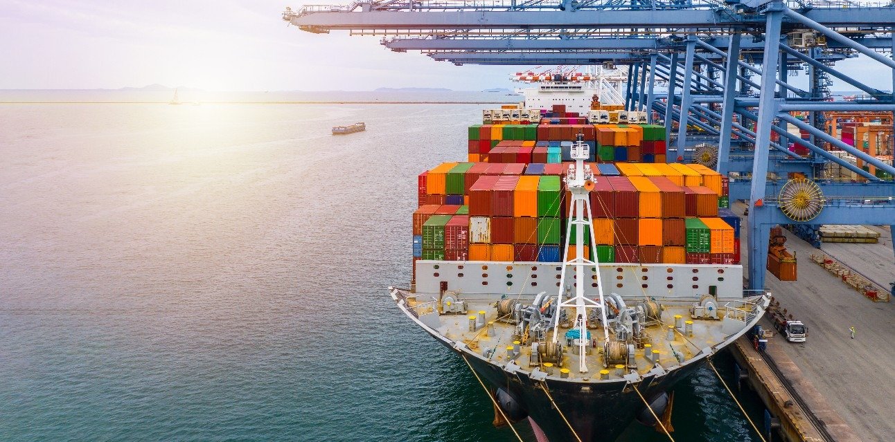 Cargo ship docked at a port with stacked colorful shipping containers on deck and blue gantry cranes overhead.