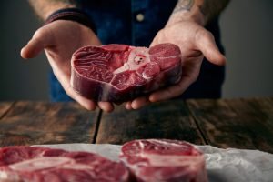 Two tattoed hands offer a piece of great meat steak with bone on camera